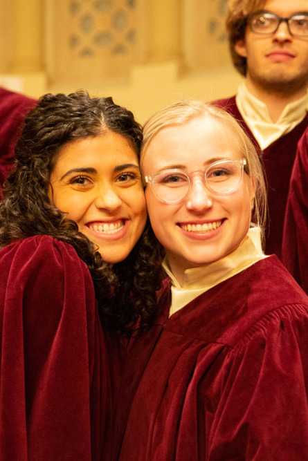 Two female students in choir performance robes hugging and smiling.