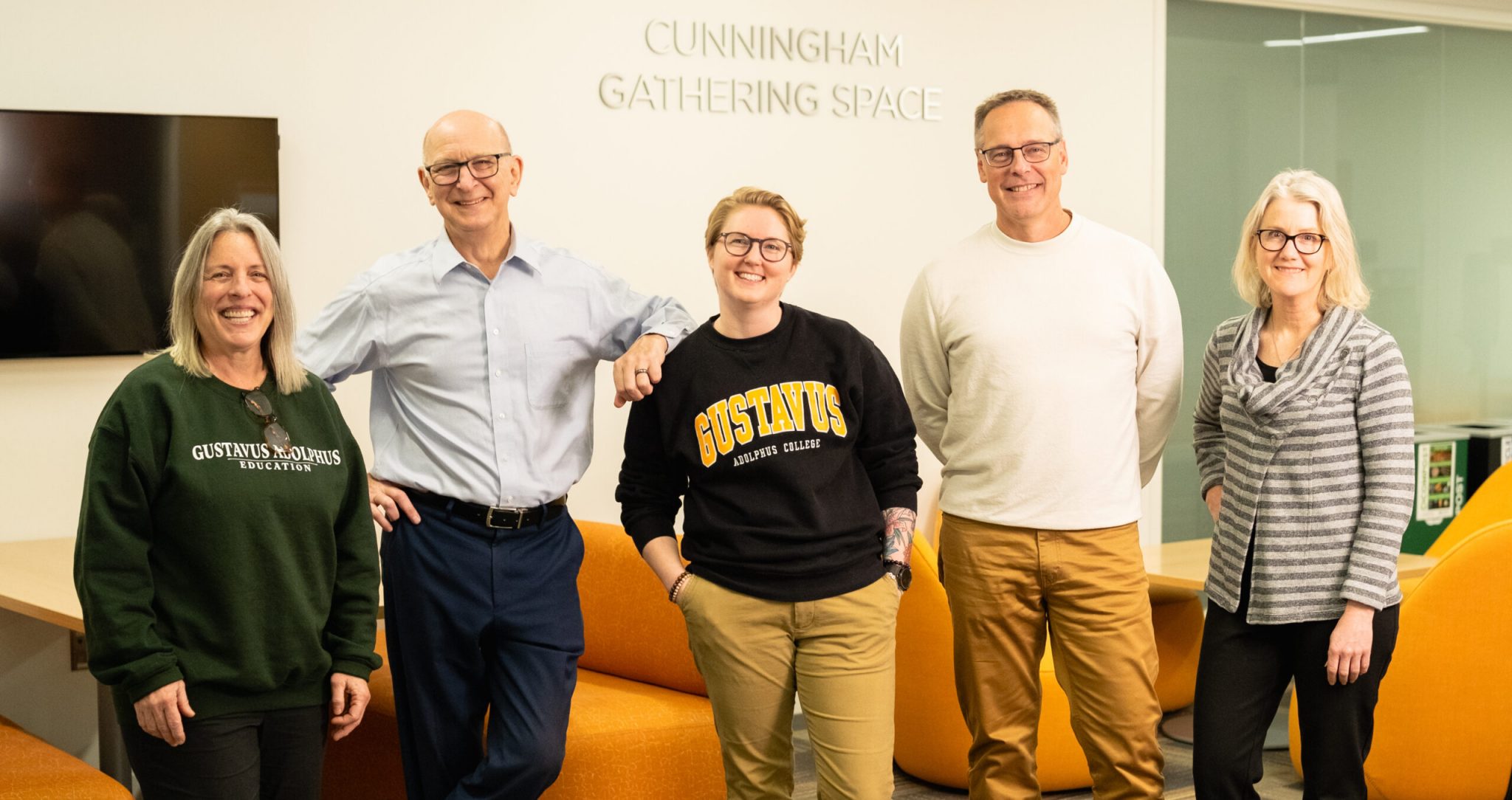 Five smiling adults pose together in a brightly lit lounge area labeled 'Cunningham Gathering Space.' They stand in front of orange furniture and a wall-mounted TV, with two of them wearing Gustavus Adolphus College apparel.