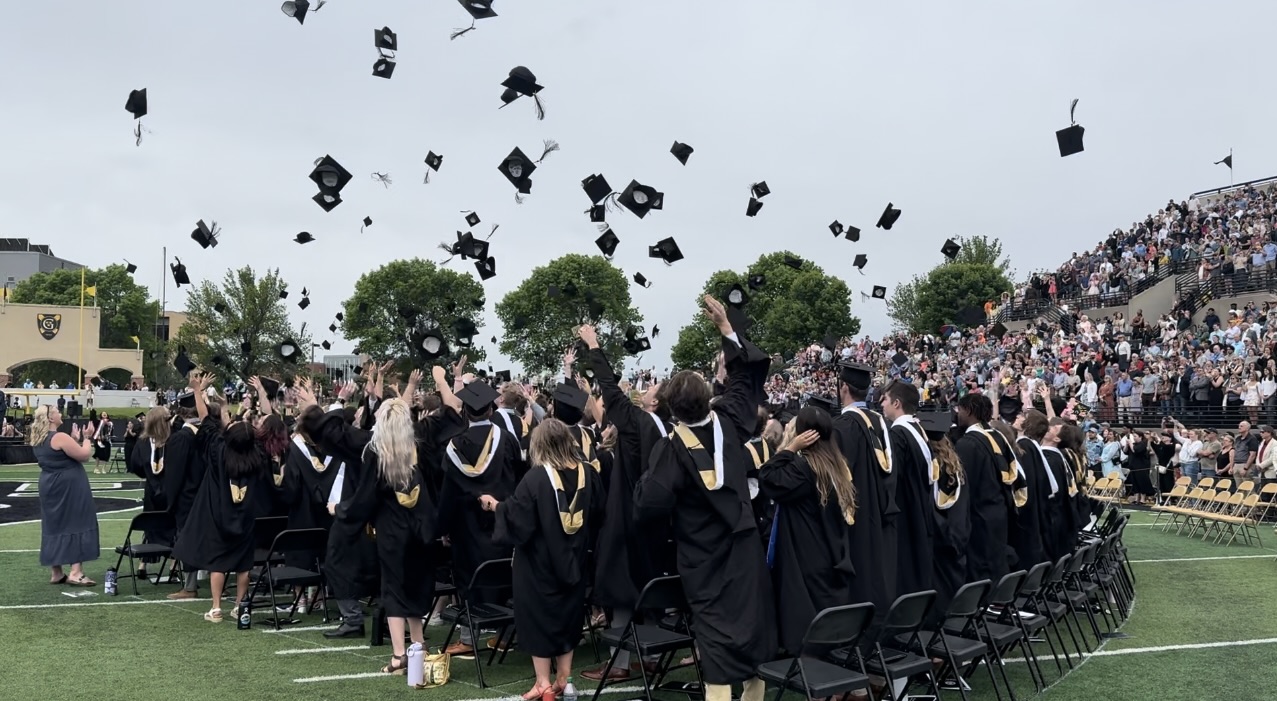 Graduates in black gowns and gold-trimmed stoles throw their caps into the air during the Gustavus Adolphus College 2024 Commencement ceremony, with cheering spectators filling the stadium stands