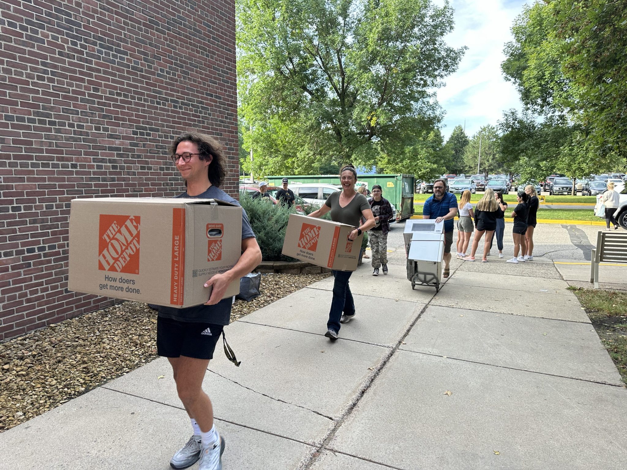 Students and families carry large boxes and carts toward a residence hall on college move-in day