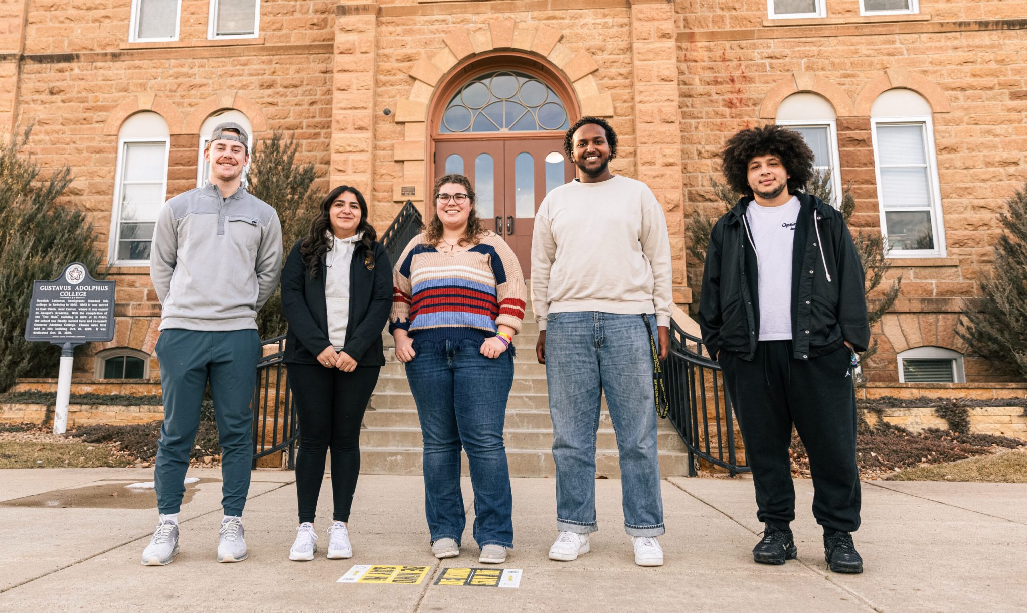 Five Gustavus Adolphus College students stand together outside a historic campus building, smiling toward the camera. A sign identifying the college is visible on the left, and informational materials are placed on the sidewalk in front of them.