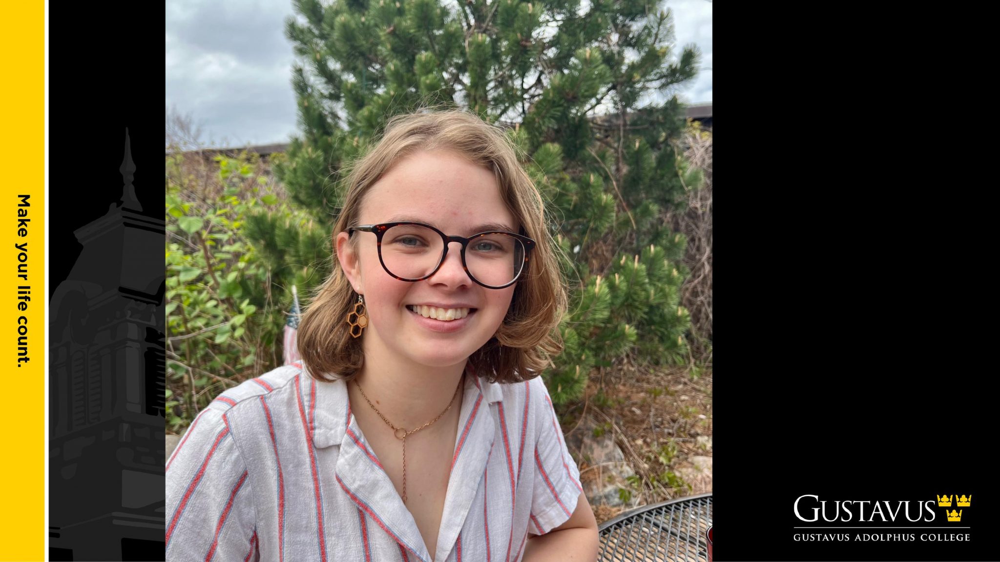 Smiling student seated outdoors with trees in the background, wearing glasses and a striped shirt, with Gustavus Adolphus College branding