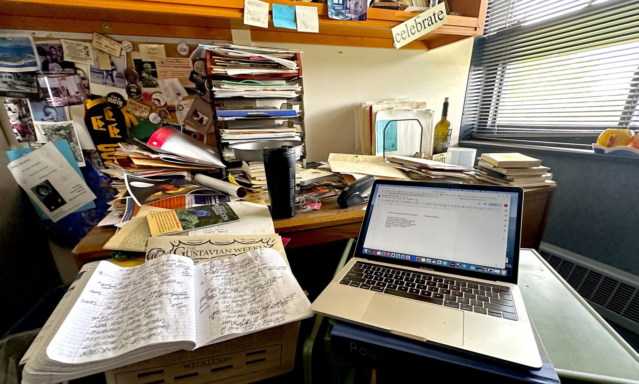 A cluttered office desk with stacks of papers, a laptop open to a document titled 'Five Questions and a Poem,' and a handwritten notebook titled 'The Gustavian Weekly'