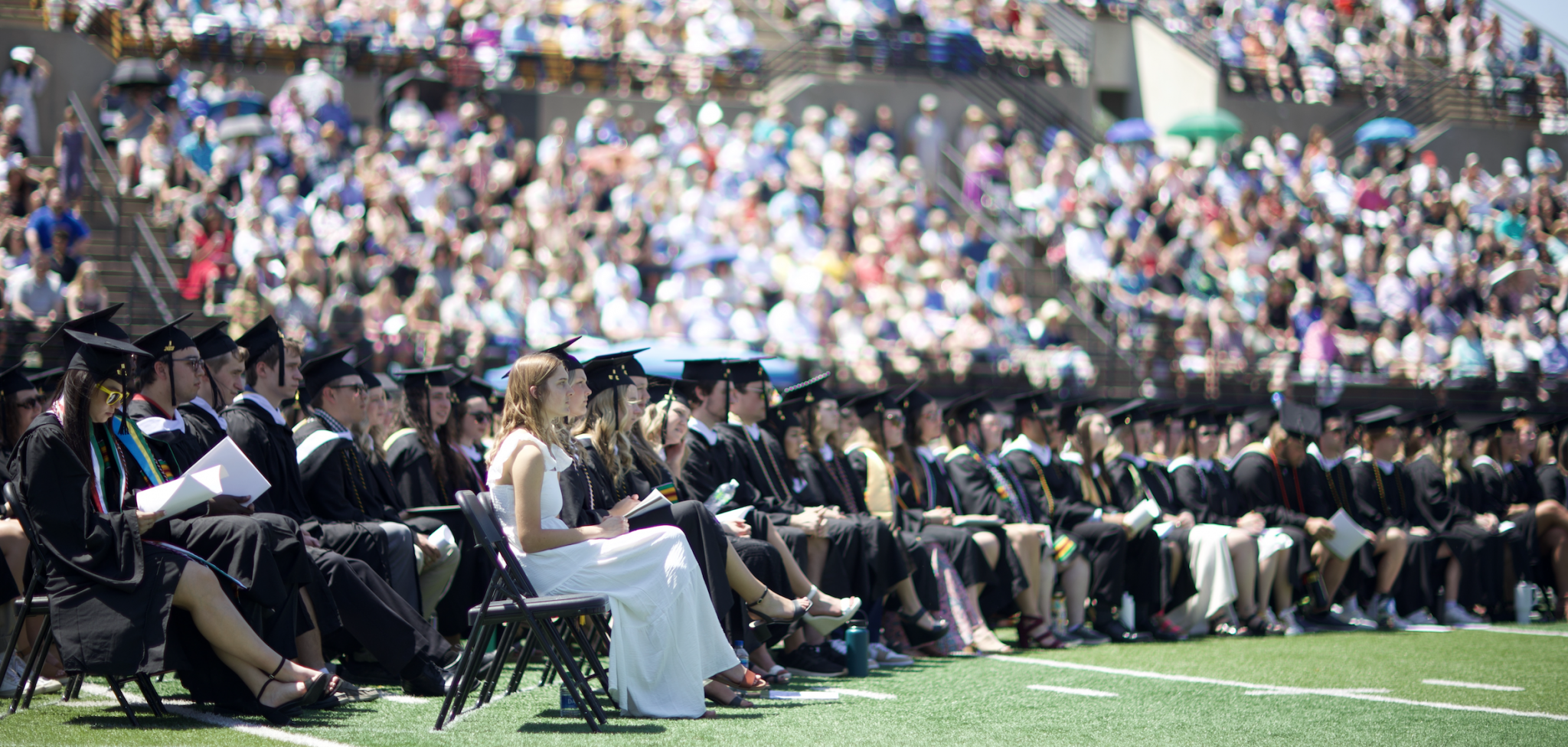 Gustavus Adolphus College graduates sit in rows on a sunny football field during the 2024 Commencement ceremony, dressed in black gowns and mortarboards, while a large crowd fills the stadium bleachers in the background.