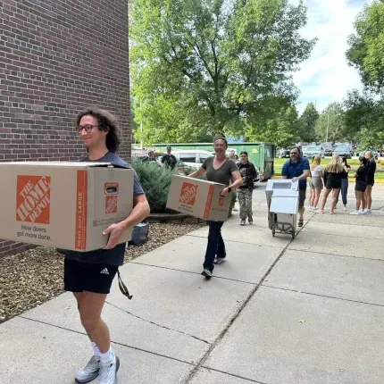 Students and families carry large boxes and carts toward a residence hall on college move-in day
