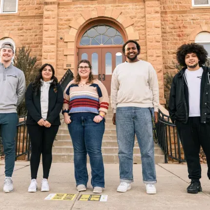 Five Gustavus Adolphus College students stand together outside a historic campus building, smiling toward the camera. A sign identifying the college is visible on the left, and informational materials are placed on the sidewalk in front of them.