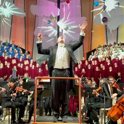 A conductor in formal attire raises his arms mid-performance in front of a large orchestra and choir. The choir is arranged in colored robes by section, blue, burgundy, and white, with dramatic lighting and abstract visuals projected behind them.
