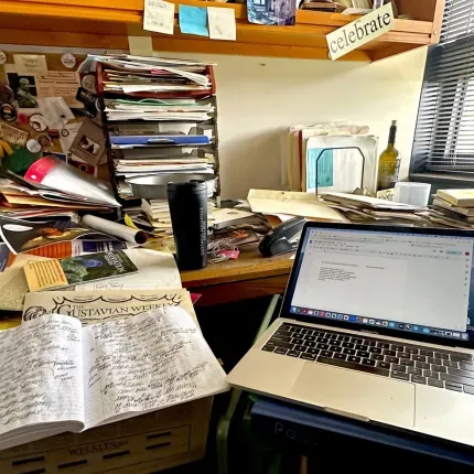 A cluttered office desk with stacks of papers, a laptop open to a document titled 'Five Questions and a Poem,' and a handwritten notebook titled 'The Gustavian Weekly'