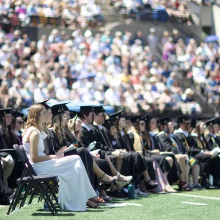 Gustavus Adolphus College graduates sit in rows on a sunny football field during the 2024 Commencement ceremony, dressed in black gowns and mortarboards, while a large crowd fills the stadium bleachers in the background.
