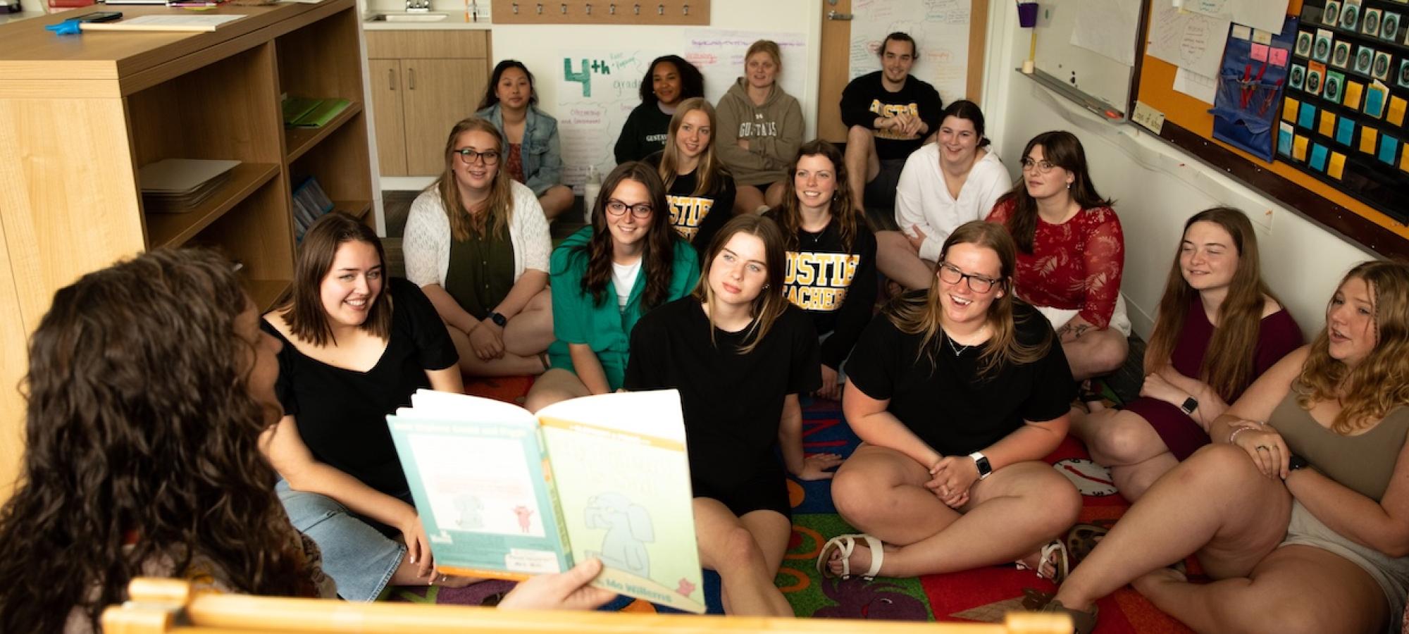 Students learning elementary education listen to a teacher as she reads a book. 