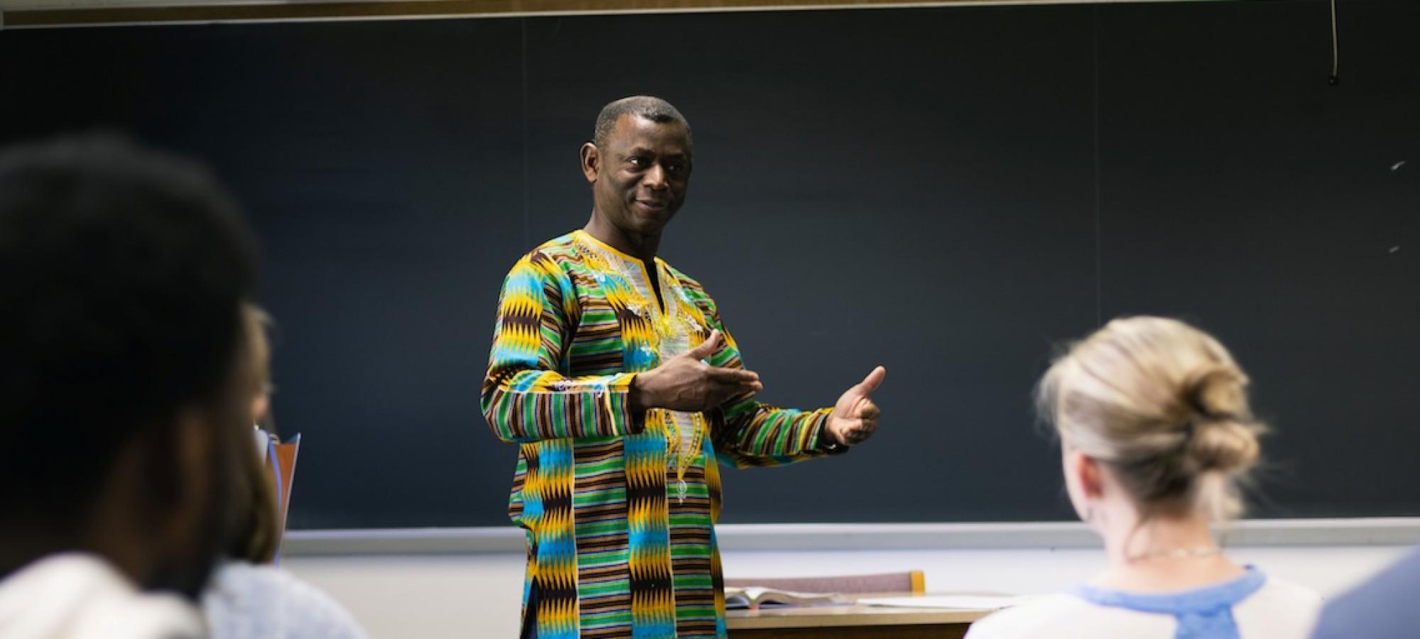 A French teacher in a colorful dashiki explains something to a class of college students. 