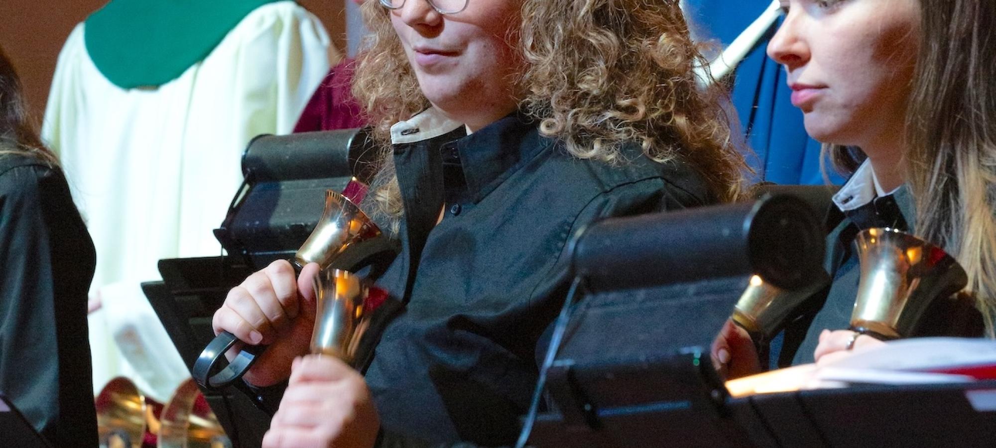 A young woman rings handbells, surrounded by fellow handbell ringers. 