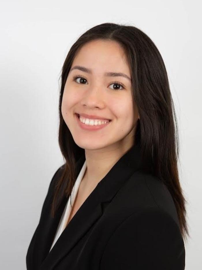 A young woman with straight dark hair smiles at the camera, wearing a black blazer and white blouse, posed in front of a plain white background