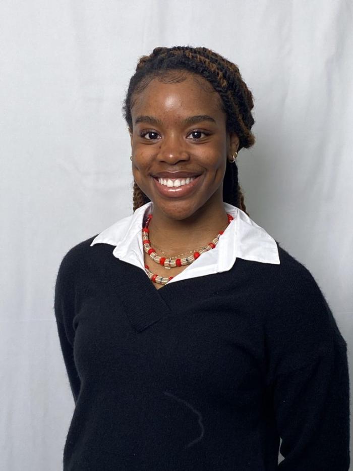 A woman smiles in front of a light gray backdrop, wearing a black V-neck sweater over a white collared shirt, accessorized with layered red and silver beaded necklaces.