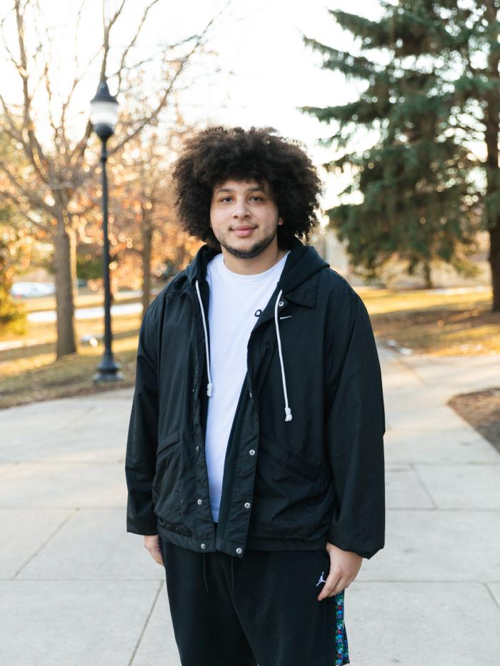 Brandon Hicks stands outdoors in the fall on a campus sidewalk, wearing a black jacket and white T-shirt.