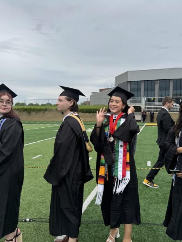 Four graduates in caps and gowns stand in a line on a football field during the Gustavus Adolphus College Commencement ceremony, smiling and chatting as they wait to proceed.