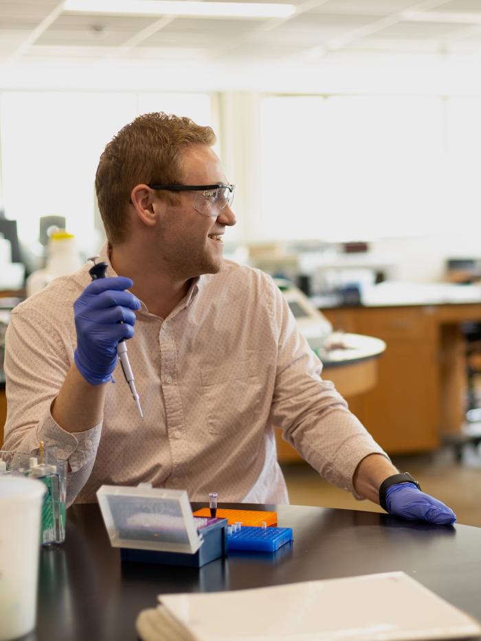 A Gustavus college student with a pipette smiles to a friend off camera. 