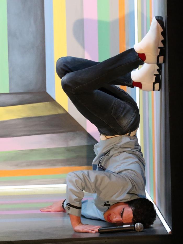 A student performer in a colorful box is upside down in a partial headstand.