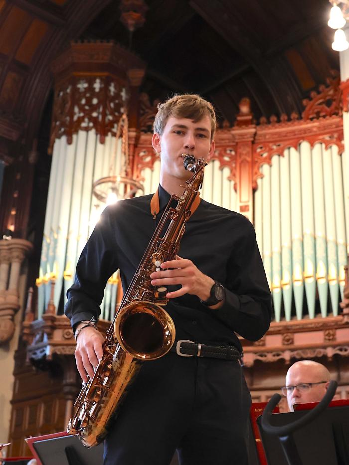 A young man blows a saxophone in front of a large organ. 